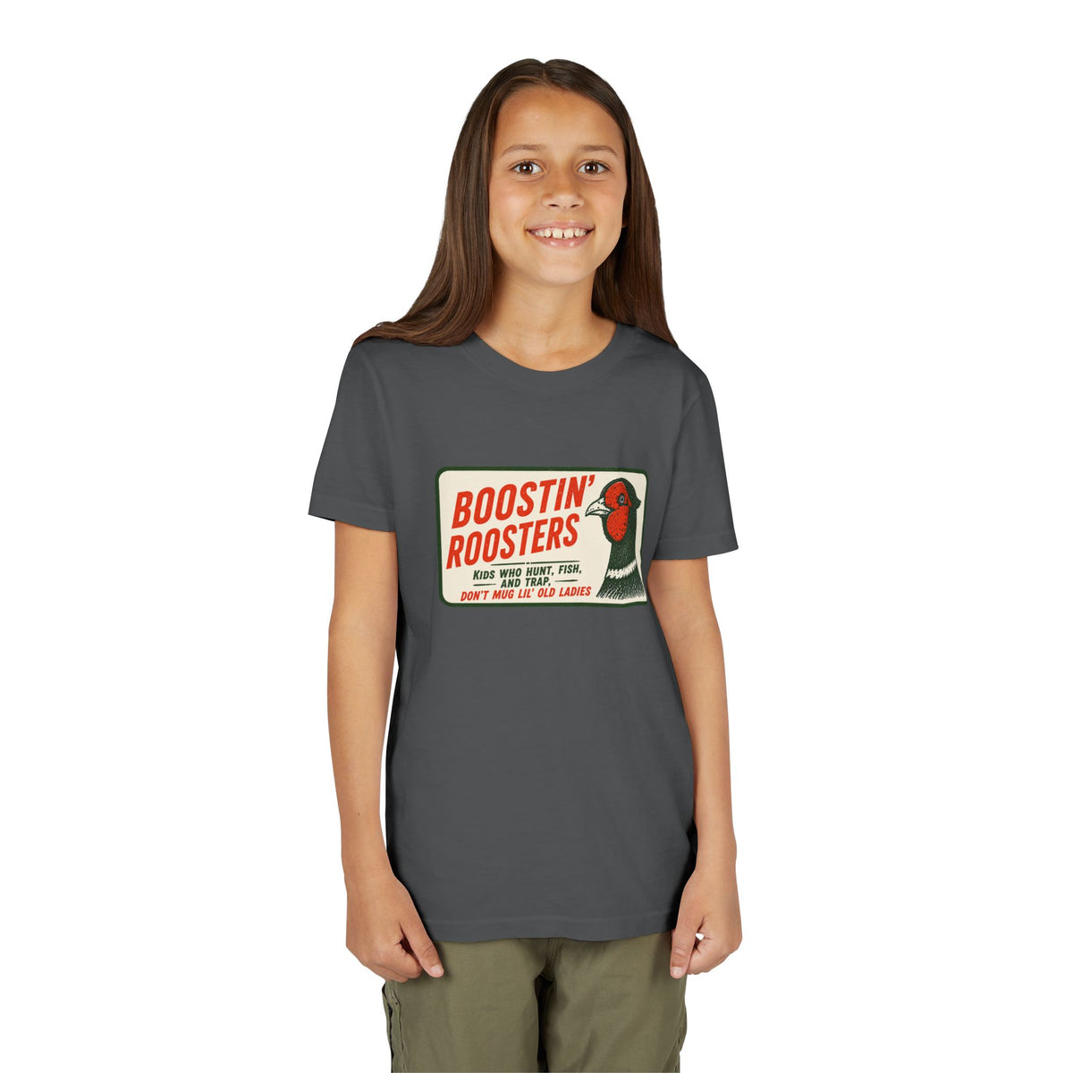 A young girl with long brown hair stands against a solid white background wearing a dark gray t-shirt with a vintage-style 'Boostin' Roosters' graphic on the chest, paired with olive green pants.