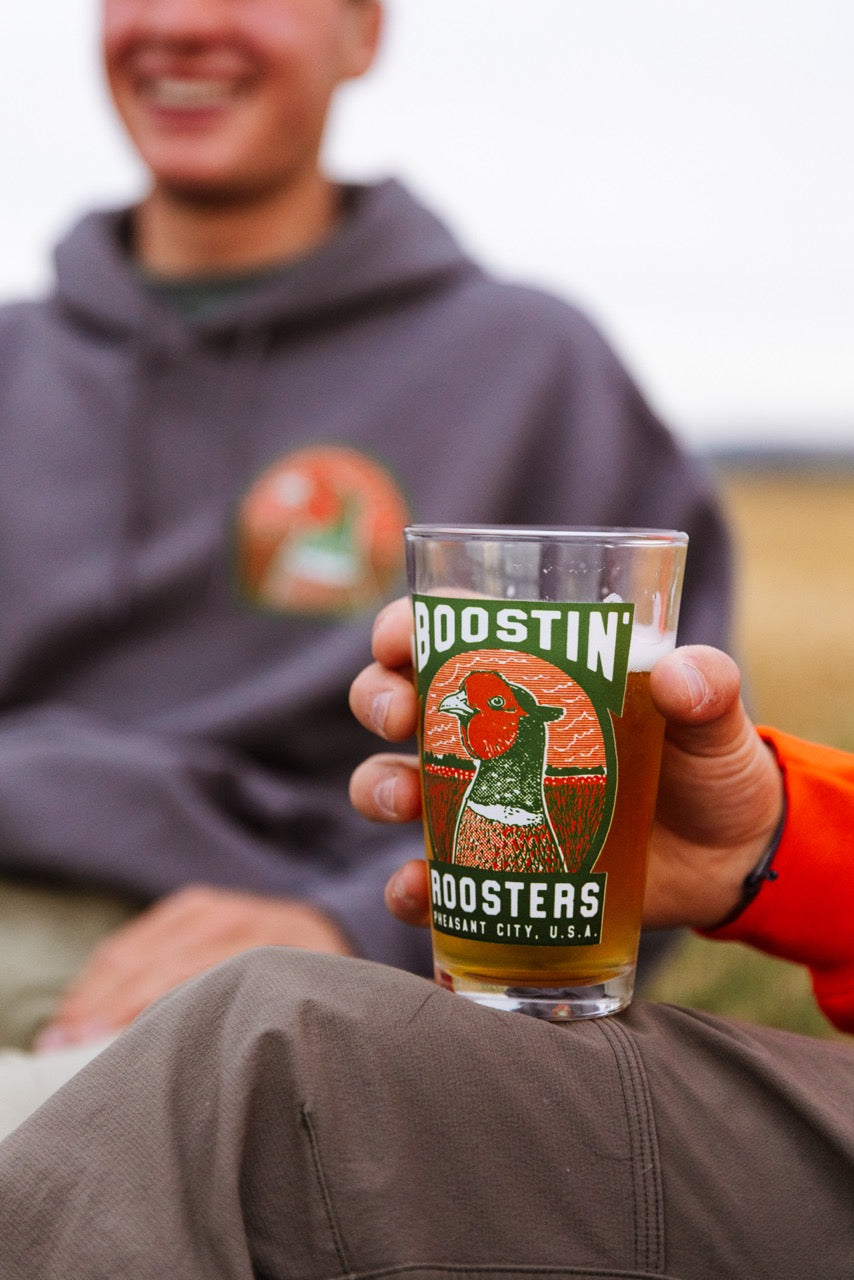 A person holding a clear pint glass with a colorful 'Boostin' Roosters' label, with another person in a gray hoodie blurred in the background outdoors.