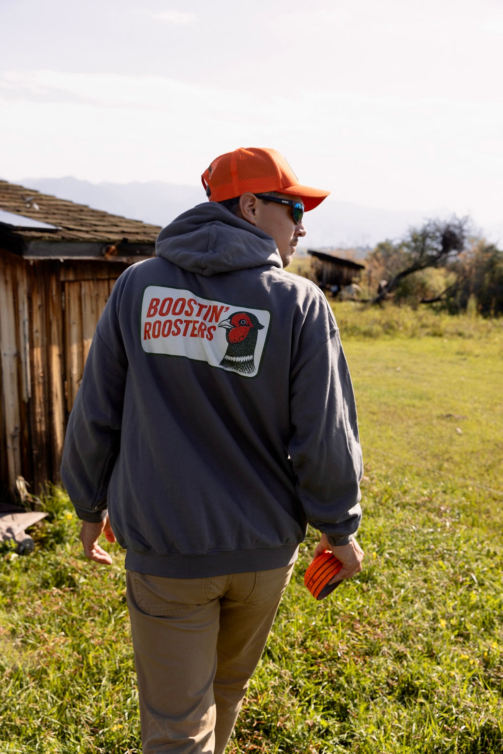 A man wearing a gray hoodie with a 'BOOSTIN' ROOSTERS' logo on the back, an orange cap, and sunglasses, walking outdoors in a grassy field near a wooden structure.