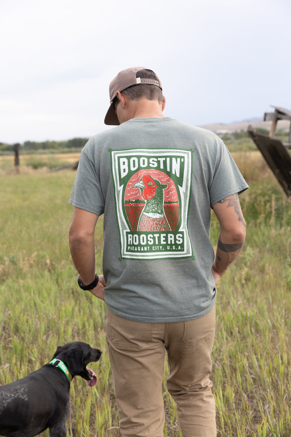 A man standing outdoors in a grassy field, viewed from behind, wearing a green t-shirt with a large 'Boostin' Roosters' graphic on the back, tan pants, and a brown cap. A black dog is standing next to him.