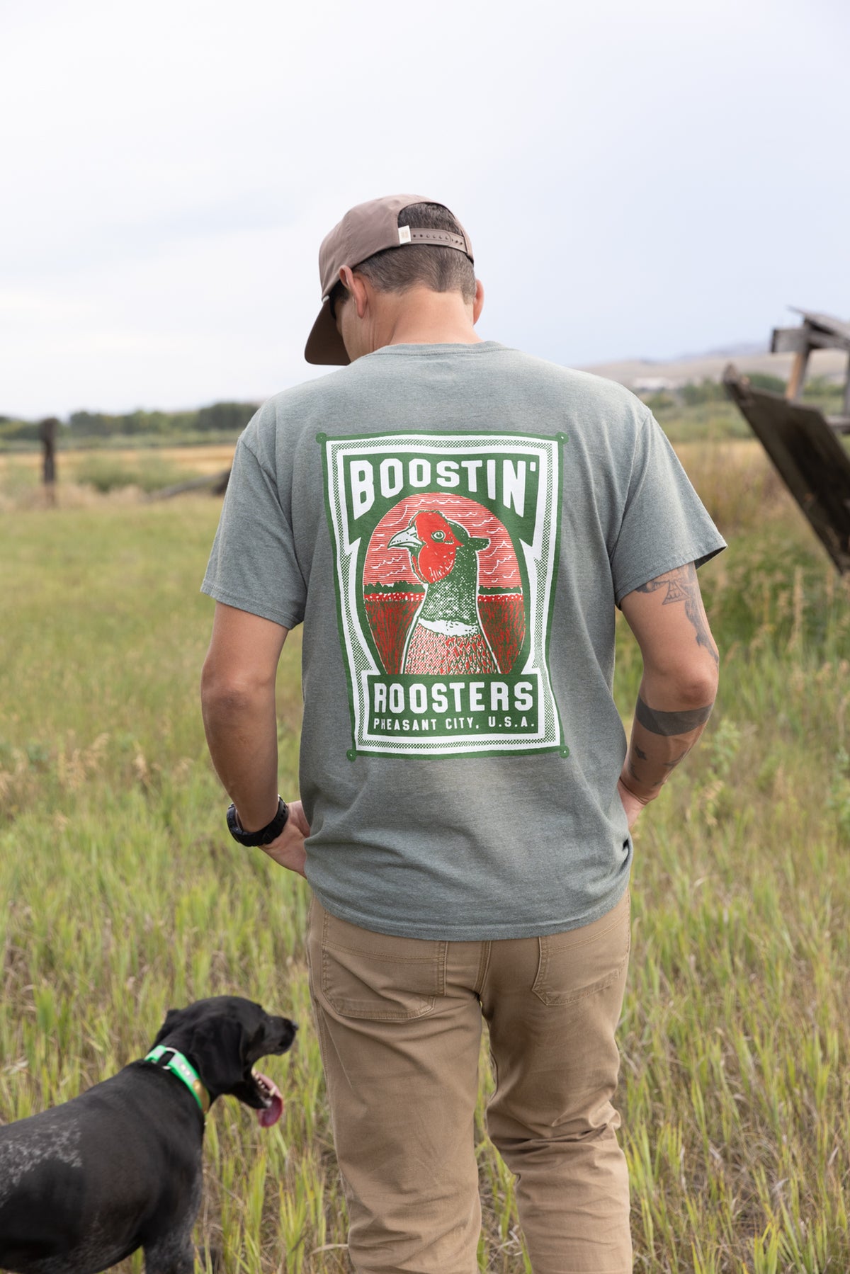 A man standing outdoors in a grassy field, viewed from behind, wearing a green t-shirt with a large 'Boostin' Roosters' graphic on the back, tan pants, and a brown cap. A black dog is standing next to him.
