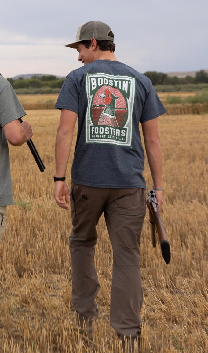 A man standing in a harvested field wearing a dark gray t-shirt with a large back graphic that reads 'BOOSTIN' ROOSTERS', brown pants, and a baseball cap, holding a shotgun.