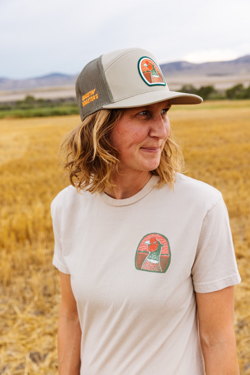 A woman standing in a golden field wearing a beige and green trucker hat with a circular pheasant head logo, and a light beige T-shirt with a matching chest graphic. The hat also features orange text "Boostin' Roosters".