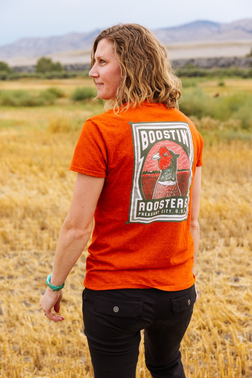 A woman standing in a dry grass field, viewed from behind, wearing a bright orange t-shirt with a large graphic print on the back that reads 'BOOSTIN ROOSTERS'.