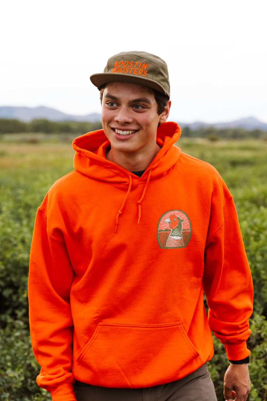 A smiling young man outdoors wearing a bright orange hoodie with a circular chest graphic of a pheasant head and an olive green cap that reads 'BOOSTIN' ROOSTERS'.