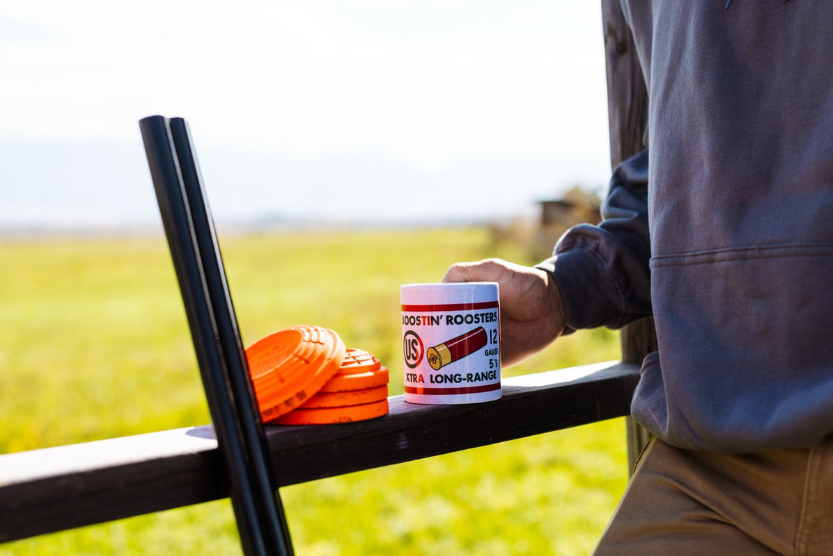 A person in a gray hoodie holds a white canister labeled 'Boostin' Booster Extra Long-Range' next to orange and black disc-like accessories on a wooden railing, with a blurred outdoor field in the background.