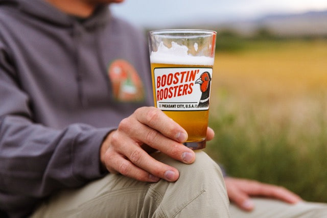A person outdoors holding a clear pint glass filled with amber beer and topped with white foam. The glass has a prominent Boostin' Boosters logo on the front.