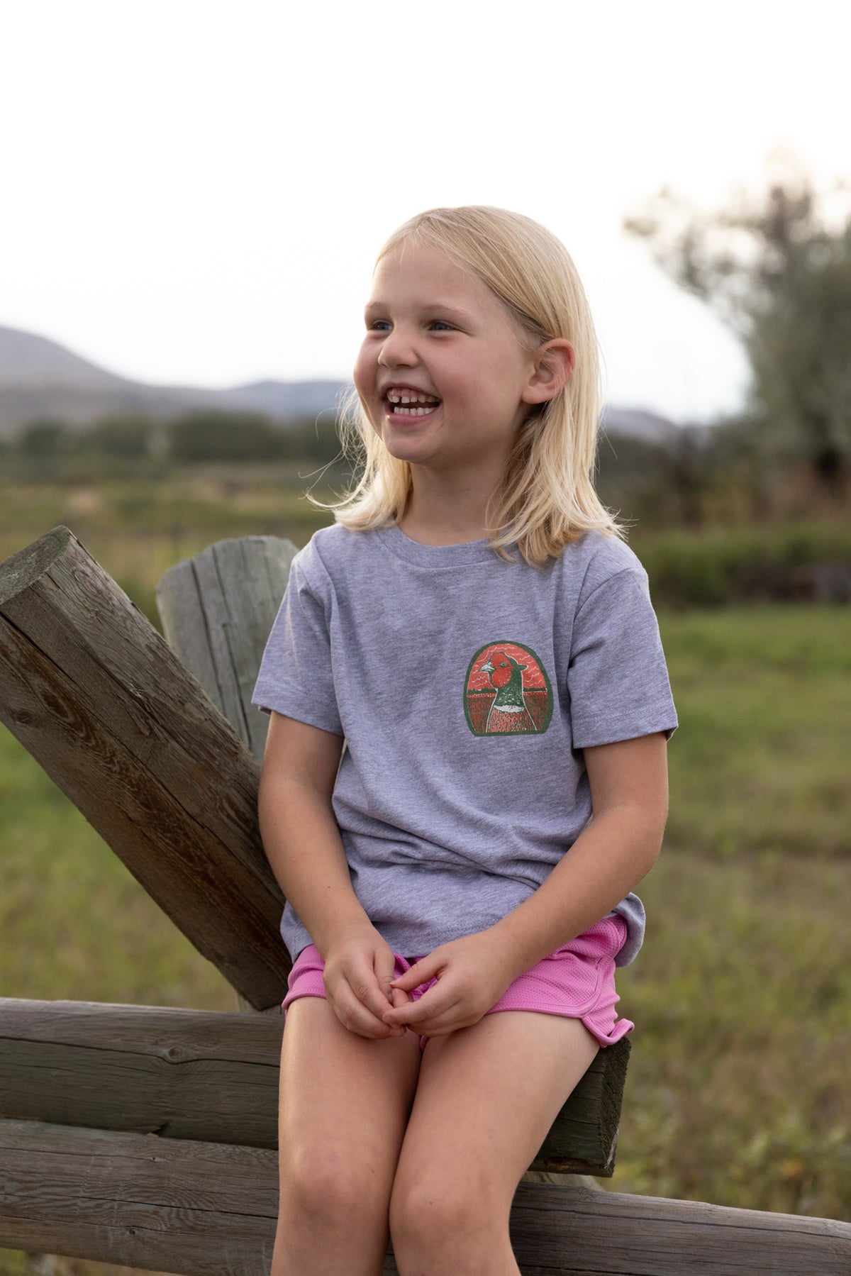 A young blonde girl sitting on a wooden fence outdoors, wearing a heather gray t-shirt with a circular pheasant head graphic on the chest and pink shorts.