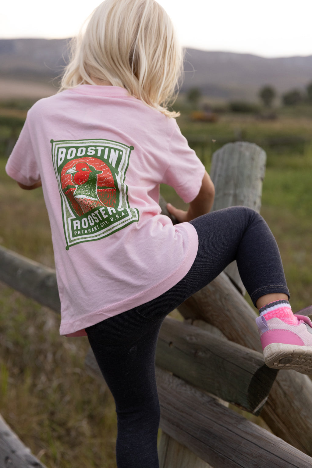 A young girl with blonde hair is seen from behind, sitting on a rustic wooden fence in an outdoor field. She is wearing a light pink t-shirt with a large "Boostin Roosters" and pheasant head graphic on the back paired with dark leggings and pink sneakers.