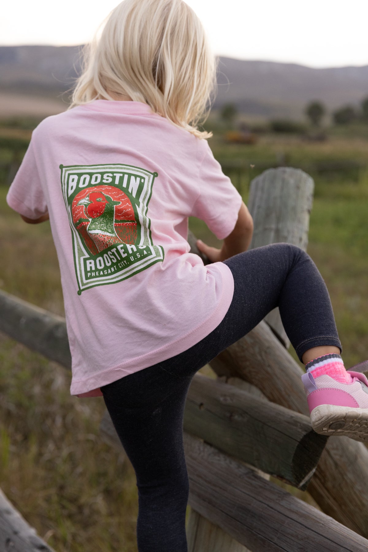 A young girl with blonde hair is seen from behind, sitting on a rustic wooden fence in an outdoor field. She is wearing a light pink t-shirt with a large "Boostin Roosters" and pheasant head graphic on the back paired with dark leggings and pink sneakers.