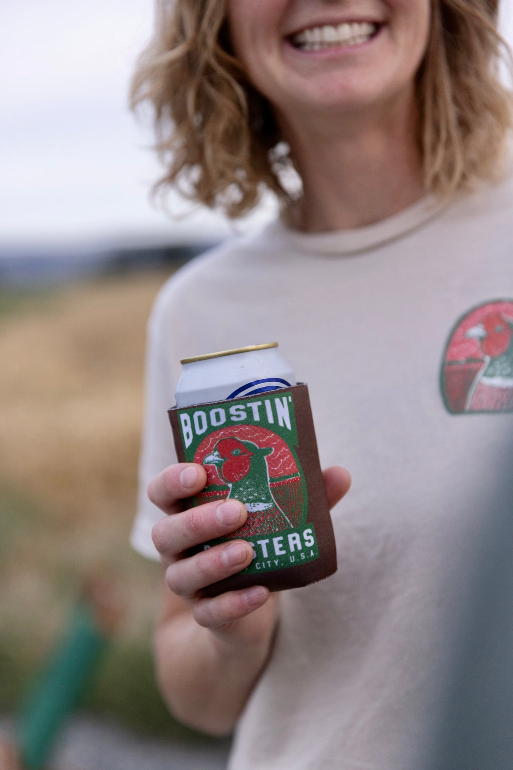 A person outdoors holding a can of Budweiser in a brown can cooler sleeve branded 'BOOSTIN''. The person is wearing a light beige t-shirt with a red Budweiser logo on the chest, and the background is a softly blurred natural outdoor setting.