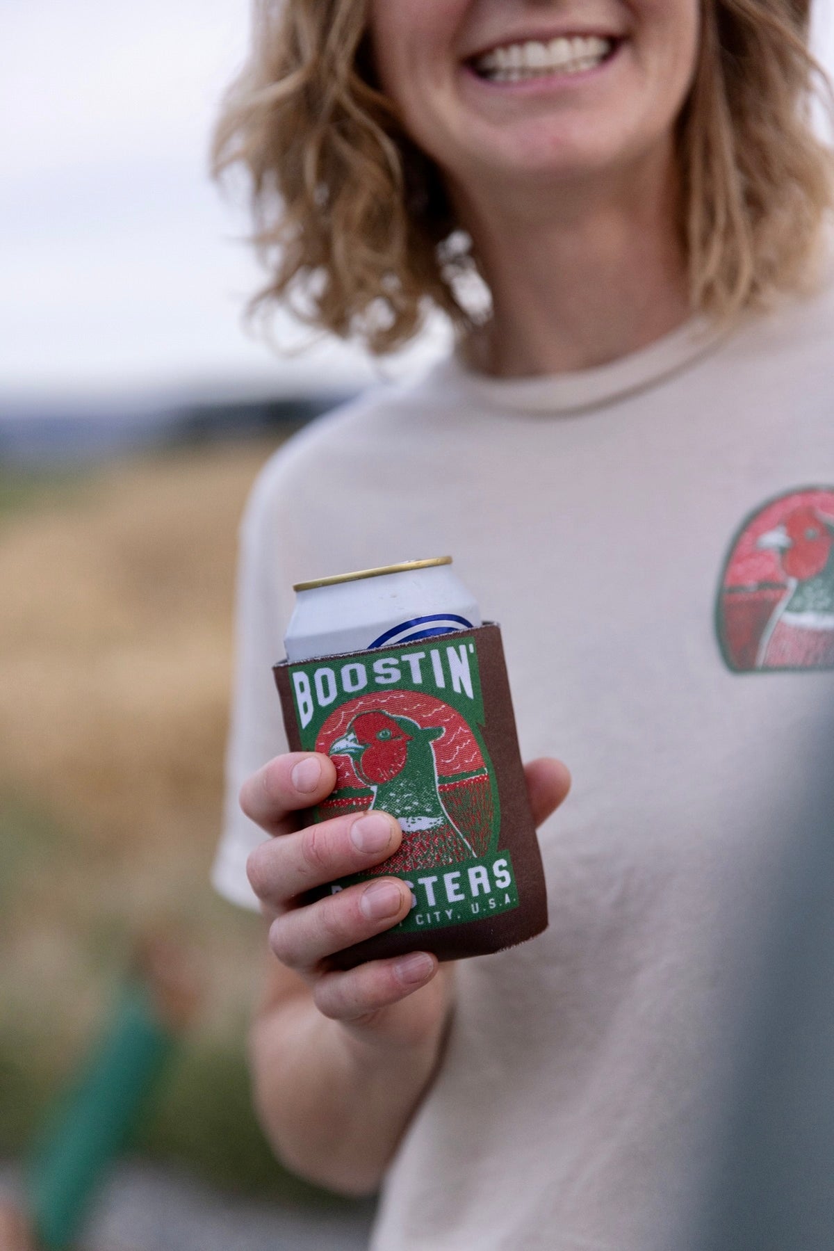 A person outdoors holding a can of Budweiser in a brown can cooler sleeve branded 'BOOSTIN''. The person is wearing a light beige t-shirt with a red Budweiser logo on the chest, and the background is a softly blurred natural outdoor setting.