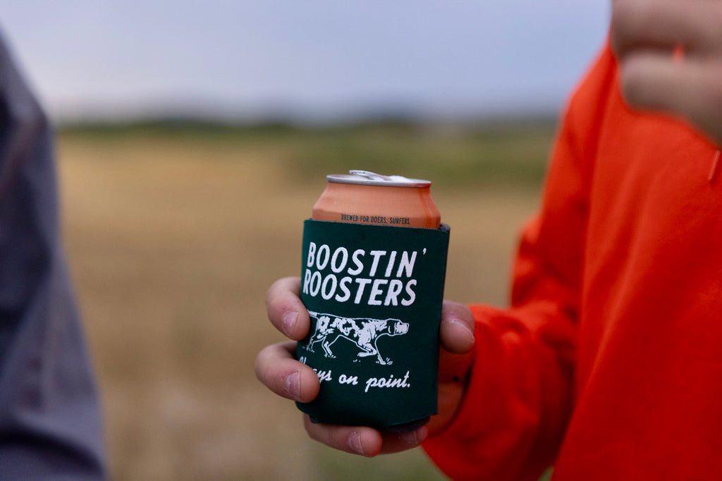 A dark green can cooler sleeve holding an orange beverage can with white text reading 'BOOSTIN' ROOSTERS' and a horse graphic, held by a person in an orange hoodie outdoors.