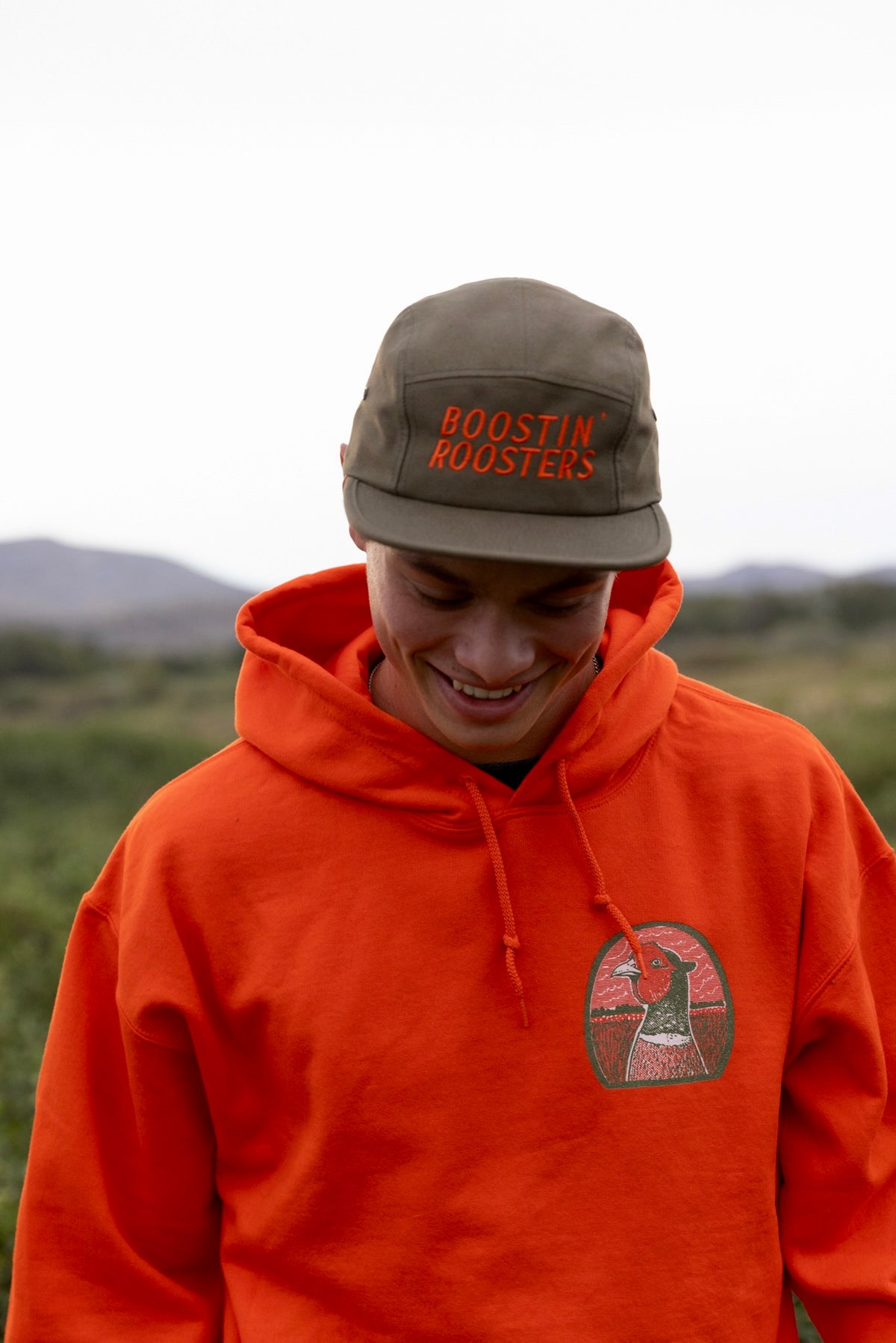 A man wearing a bright orange hoodie and a cap with 'BOOSTIN ROOSTERS' embroidered in orange, standing outdoors against a soft-focus natural background.