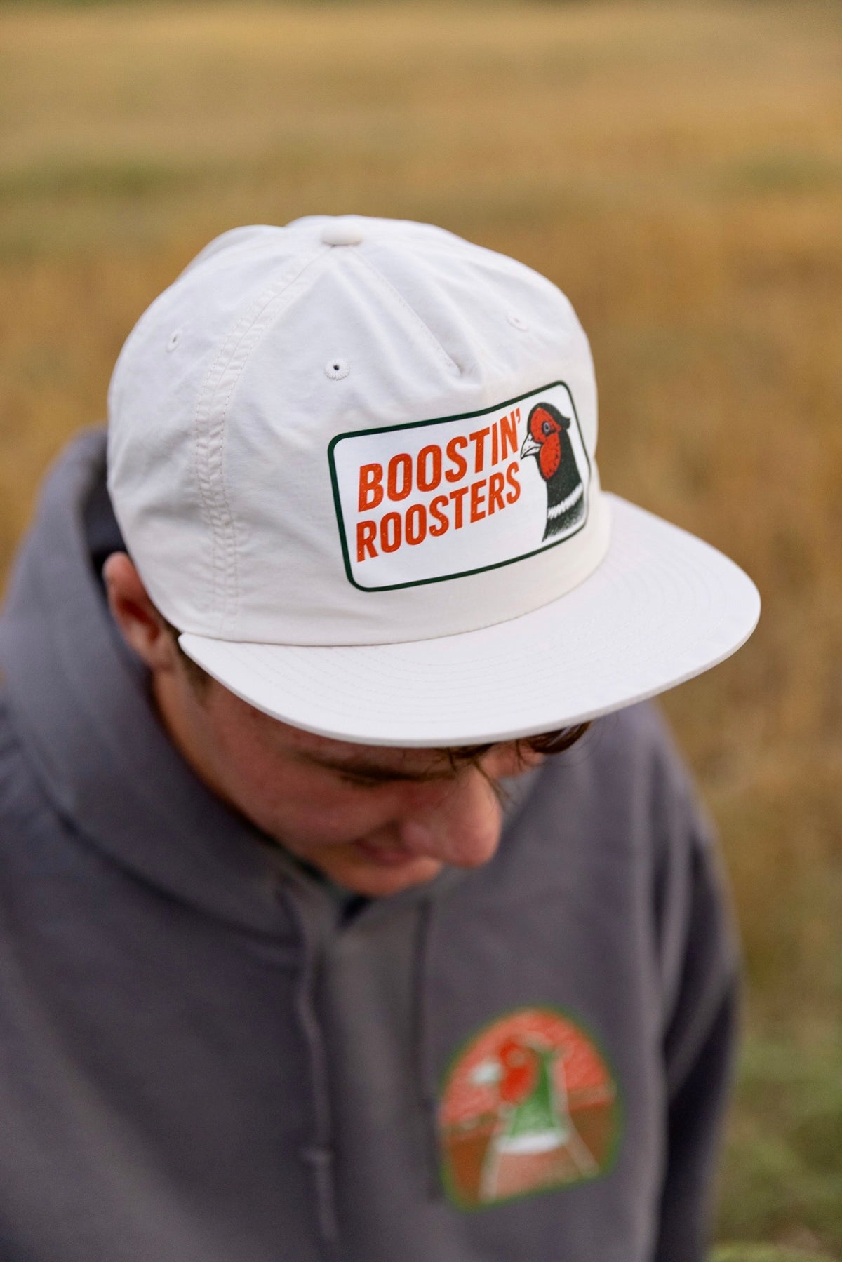 A person wearing a white flat-brim cap with a 'BOOSTIN' ROOSTERS' logo patch on the front, photographed outdoors against a blurred tan field background.