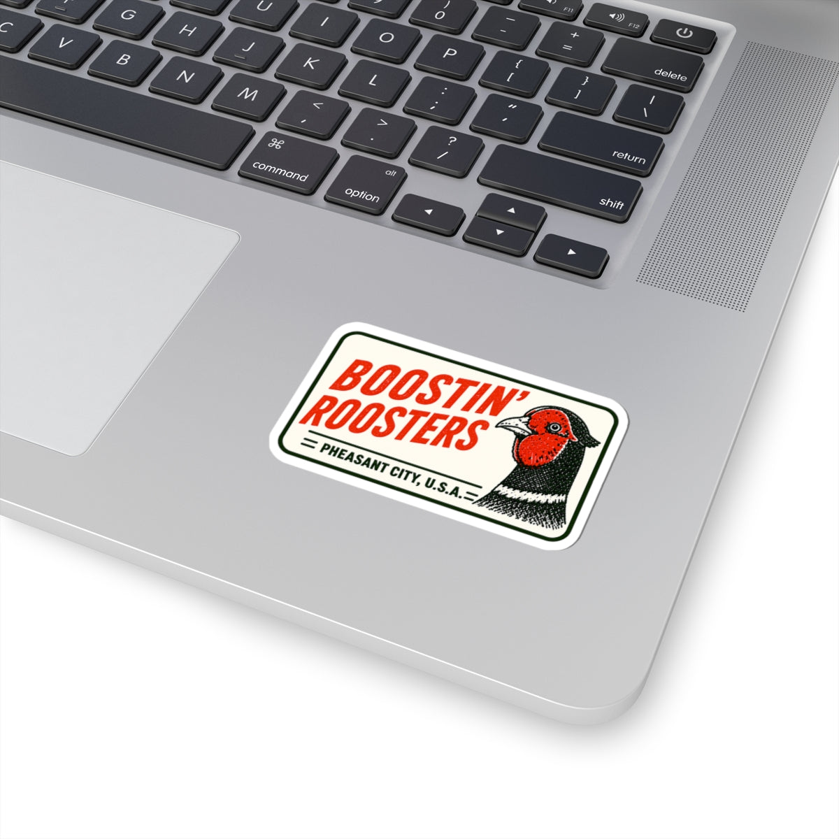 A rectangular Boostin' Roosters sticker applied to the palm rest of a silver laptop. The sticker features red text, a black and red rooster illustration, and 'Pheasant City, USA
