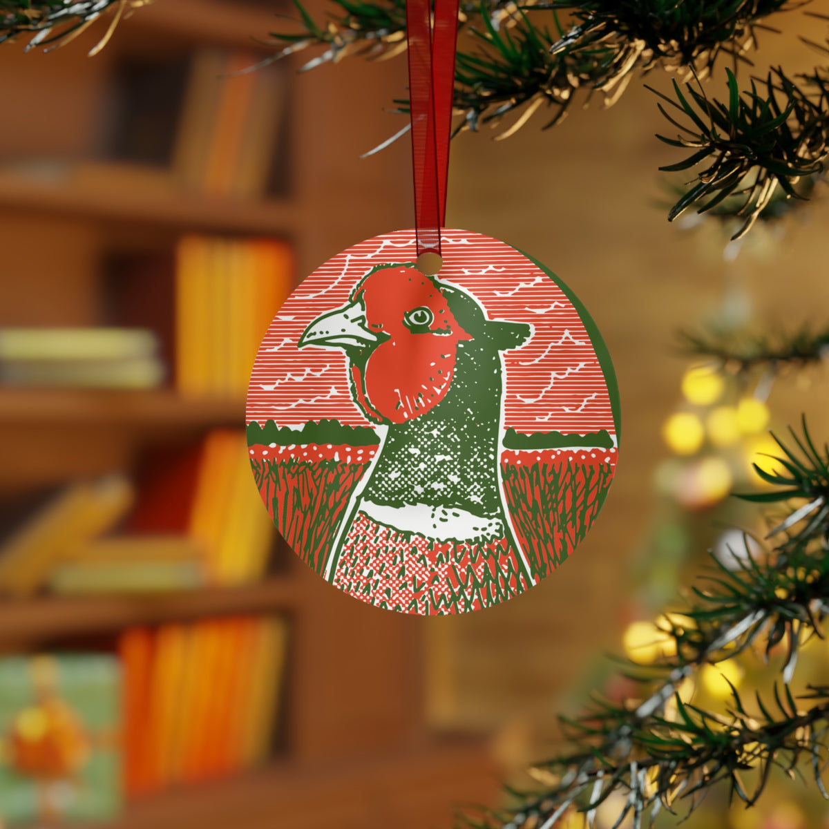 A round Christmas ornament featuring a stylized pheasant bird in red, green, and white, hanging from a red ribbon on a Christmas tree branch with a blurred indoor background.