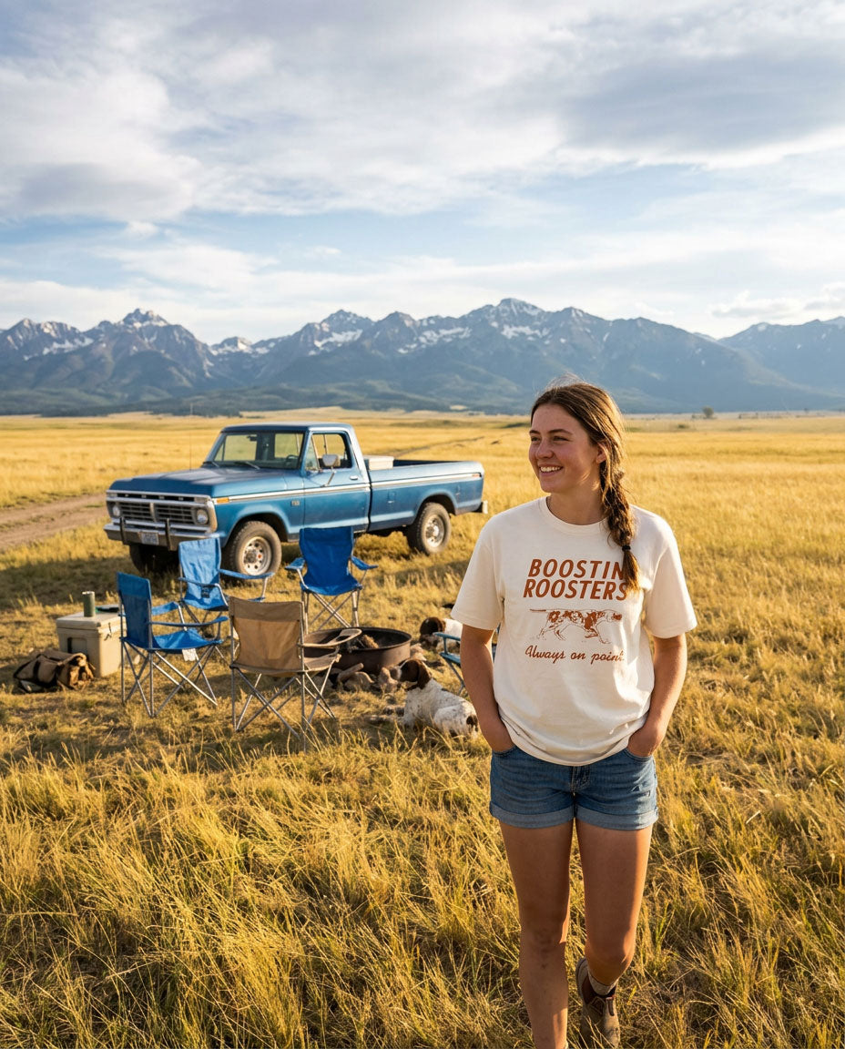A woman stands in a grassy field wearing a cream graphic t-shirt that reads 'BOOSTIN ROOSTERS' and denim shorts, with a blue pickup truck and camping chairs in the background against a mountain range.