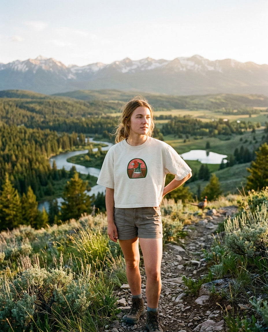 A woman stands on a mountain trail wearing a cream-colored Boostin' Roosters graphic t-shirt and brown shorts, with a winding river and snow-capped mountains in the background.